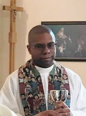 Portrait of Bishop Theogene wearing a white alb and a mulitcolered stole.  there is a crucifix and a framed picture in the background